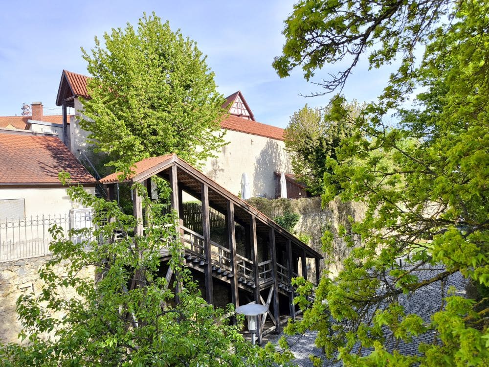 Eine überdachte Holztreppe führt hinauf zu beigen Gebäuden mit roten Ziegeldächern in Nördlingen, umgeben von grünen Bäumen und Laub an einem sonnigen Tag.