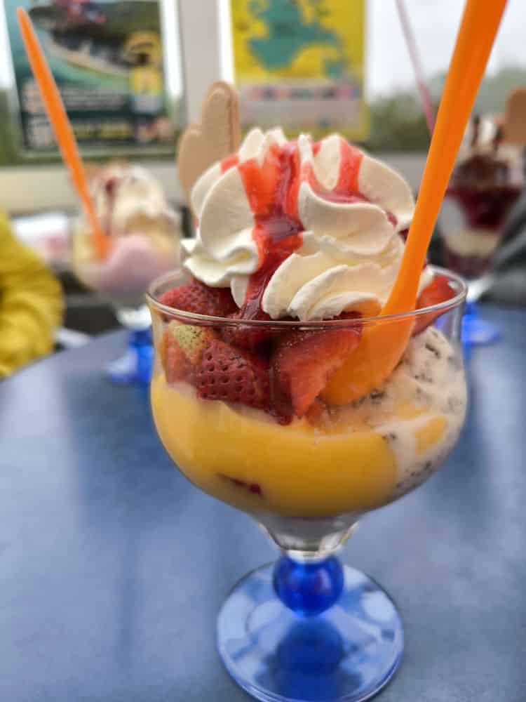 Ein gläserner Dessertbecher gefüllt mit Vanilleeis, Erdbeeren, Vanillepudding, Schlagsahne und Erdbeersauce, serviert mit einem orangenen Löffel und einer Waffel auf einem blauen Tisch - perfekt zum Genießen im Lieblingsrestaurant Rügen.