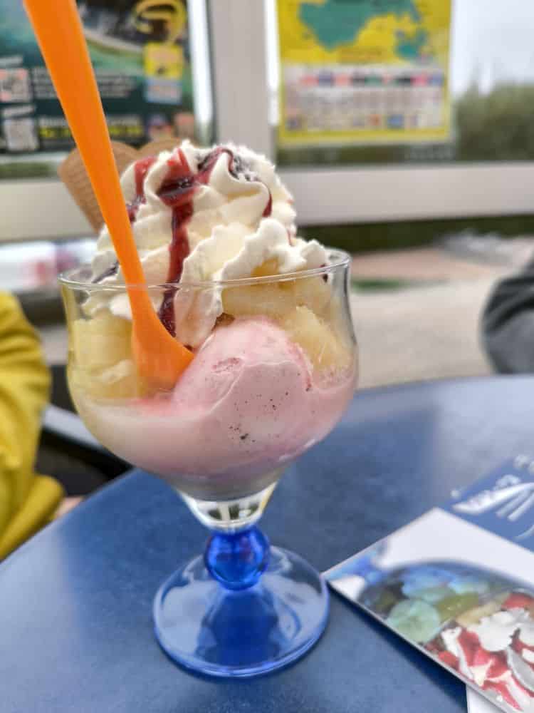 Ein gläserner Dessertbecher mit rosa-weißem Eis, Schlagsahne, Bananenscheiben und einem Waffelstück wird mit einem orangefarbenen Löffel auf einem blauen Tisch serviert - perfekt zum Genießen in einem der Lieblingsrestaurants Rügen.