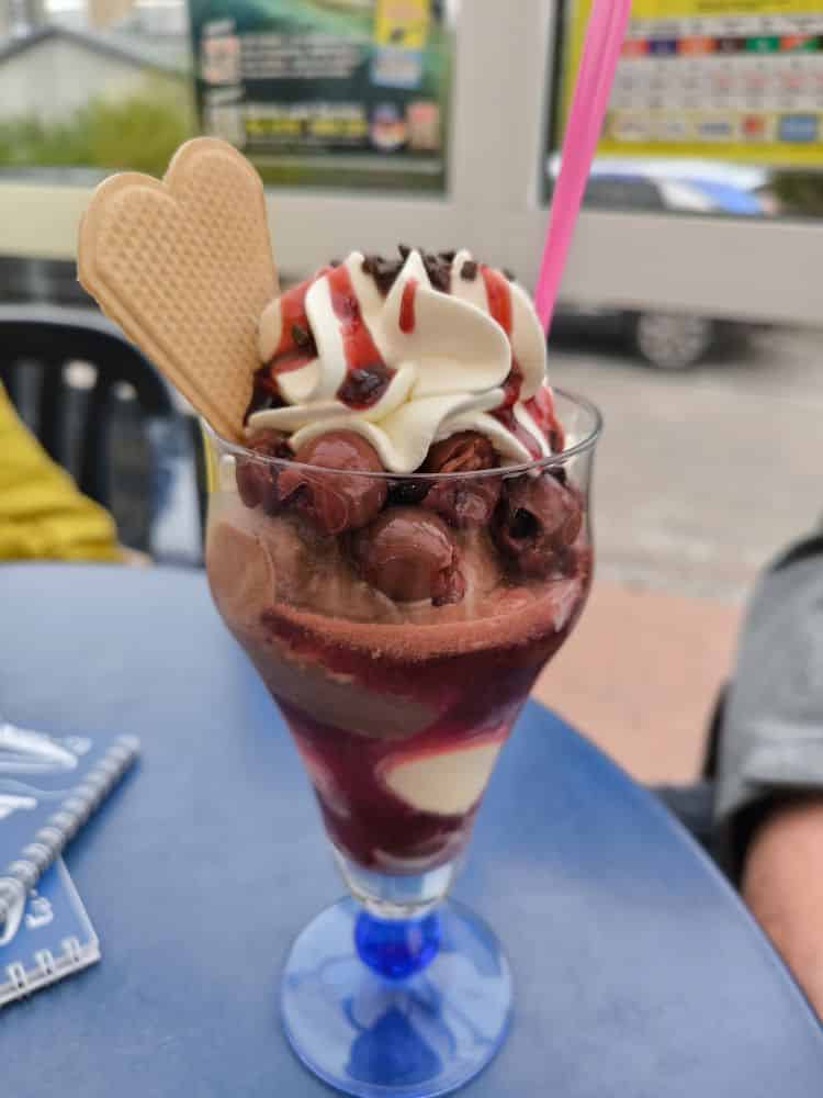 Auf einem blauen Tisch steht ein hohes Glas mit Schokoladen- und Vanilleeis, das mit Schlagsahne, Kirschen, Schokoladenstückchen, einer herzförmigen Waffel und einem rosa Strohhalm gefüllt ist - perfekt für den Genuss in den Lieblingsrestaurants auf der Insel Rügen.