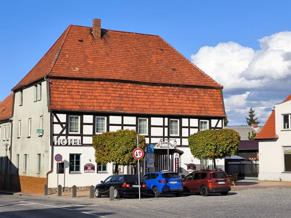 An einer Straßenecke in Neubukow steht ein traditionelles Hotelgebäude in Fachwerkbauweise mit rotem Ziegeldach. Mehrere Autos sind davor geparkt, und zwei beschnittene Bäume flankieren den Hoteleingang unter einem blauen Wolkenhimmel.