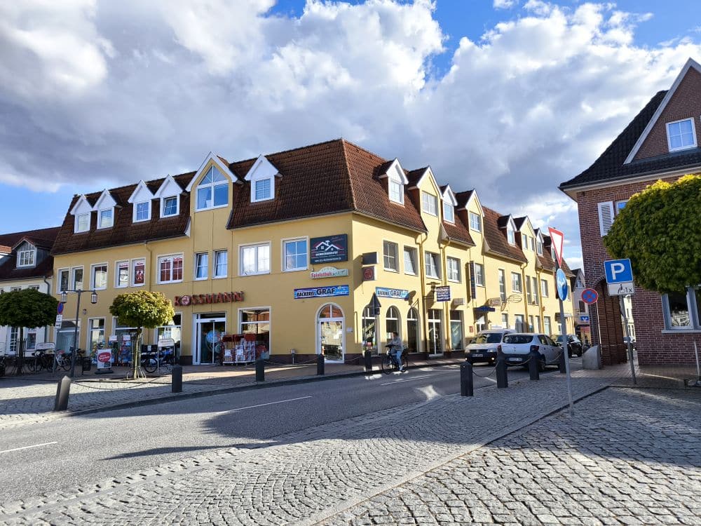 Eine Straßenansicht in Neubukow zeigt ein gelbes Eckhaus mit Geschäften im Erdgeschoss, Fahrräder, die vor dem Haus geparkt sind, Menschen, die auf dem Kopfsteinpflaster flanieren, und einen teilweise bewölkten Himmel über sich.