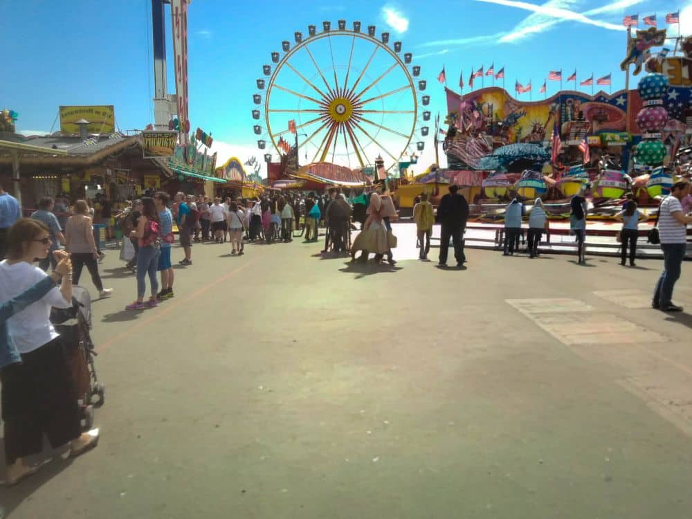 Ein lebhafter Rummelplatz mit einem Riesenrad, bunten Fahrgeschäften und Menschen, die unter einem strahlend blauen Himmel herumlaufen. Die Oktoberfestdekoration sorgt für festlichen Charme, während die Besucher Fotos machen, sich in Gruppen versammeln oder durch den belebten Bereich schlendern.