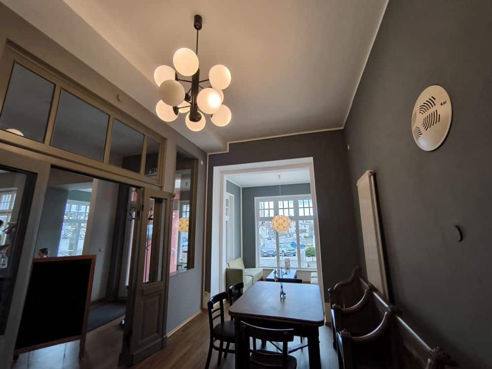 Ein moderner Essbereich mit grauen Wänden, einem Holztisch und Stühlen sowie einem einzigartigen runden Kronleuchter. Große Fenster lassen natürliches Licht herein und bieten einen Blick, der an das Restaurant Rügen auf der malerischen Insel Rügen erinnert.