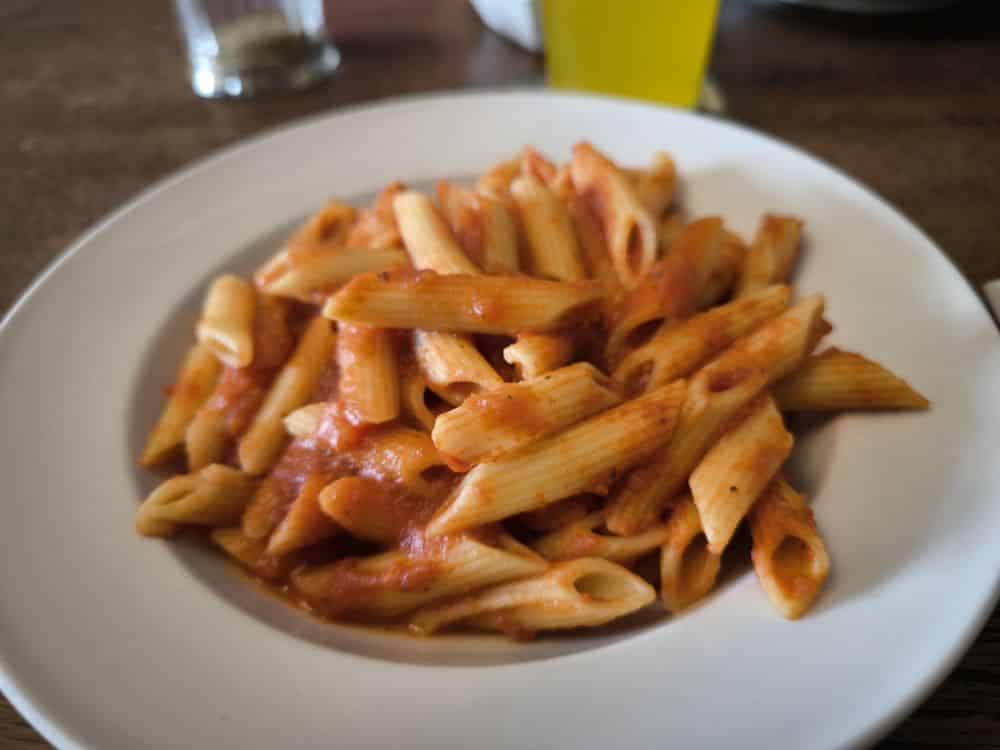 Ein weißer Teller mit Penne-Nudeln und Tomatensoße steht auf einem braunen Tisch. Im Hintergrund sind teilweise ein gelbes Glas und ein klares Glas zu sehen - perfekt für ein Essen in einem der gemütlichen Restaurants auf Rügen.