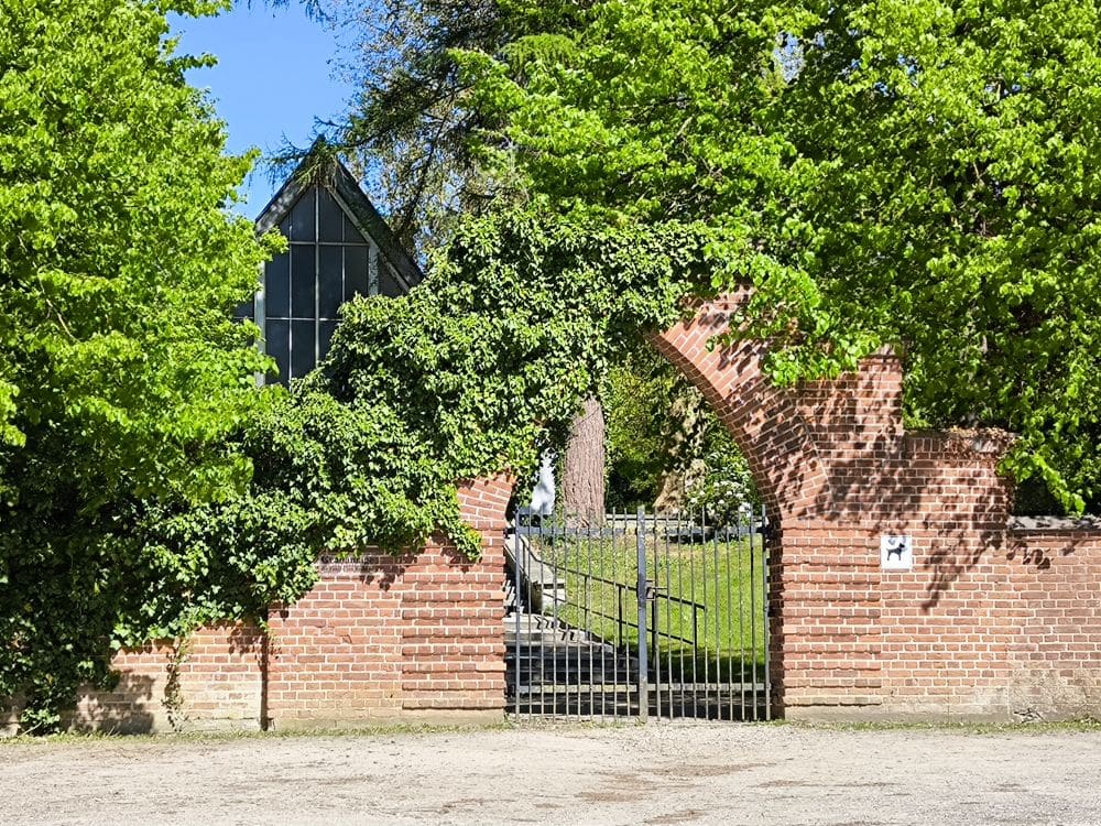 Eine Backsteinmauer mit einem gewölbten Eisentor, das teilweise von grünem Efeu bedeckt ist, umgeben von üppigen Bäumen, erinnert an den Charme von Klütz an der Ostseeküste. Durch das Tor hindurch sind im Hintergrund ein Hof und ein Gebäude mit einer Glasfassade zu sehen.