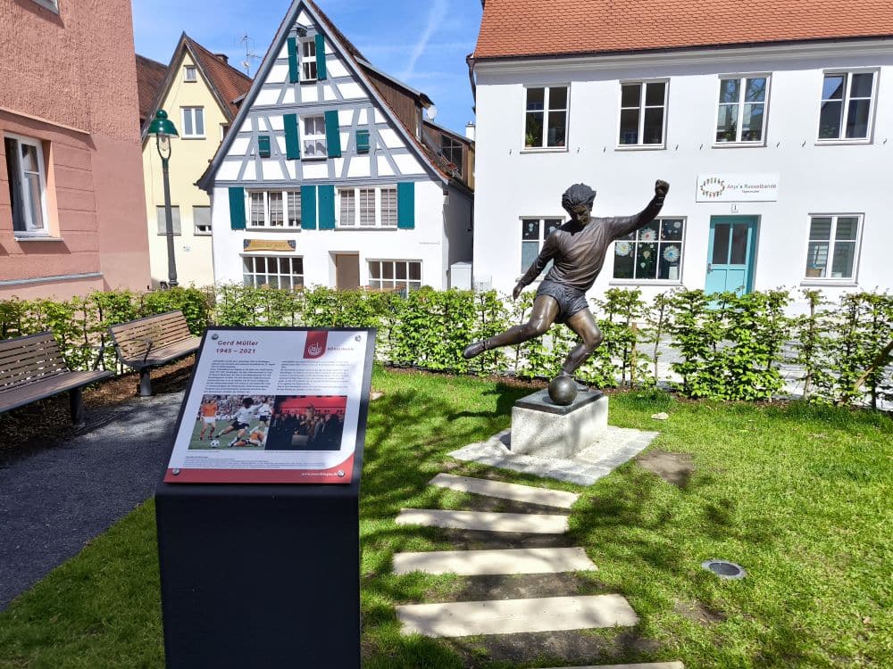 Die Bronzestatue eines Fußballspielers, der einen Ball kickt, steht auf einem Sockel in einem kleinen Park mit grünem Rasen in Nördlingen. In der Nähe laden eine Informationstafel und Bänke zum Ausruhen ein, während traditionelle europäische Häuser eine reizvolle Kulisse bilden.