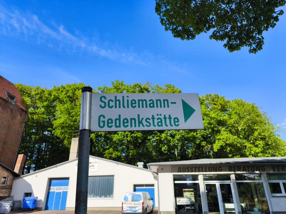 Ein Straßenschild mit der Aufschrift Schliemann-Gedenkstätte und einem nach rechts zeigenden Pfeil steht in Neubukow vor einem Gebäude, Bäumen und einem strahlend blauen Himmel.