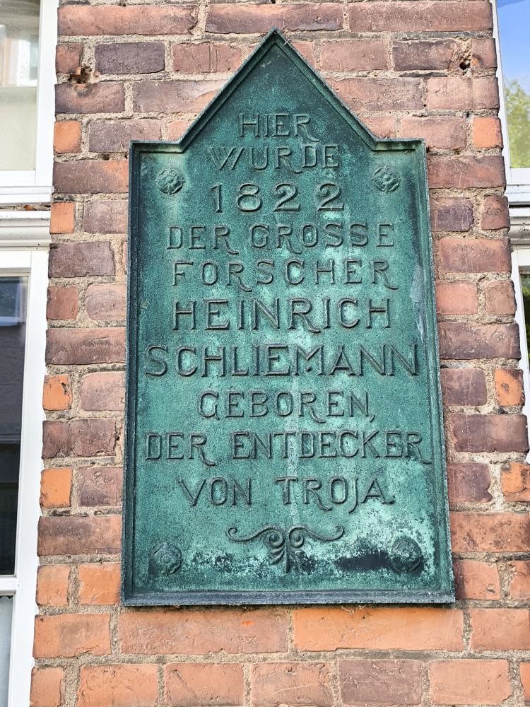 Auf einer grünen Metalltafel an einer Backsteinmauer in Neubukow steht auf Deutsch: "Hier wurde 1822 der große Forscher Heinrich Schliemann geboren, der Entdecker von Troja", womit Schliemanns Geburtshaus gekennzeichnet ist.