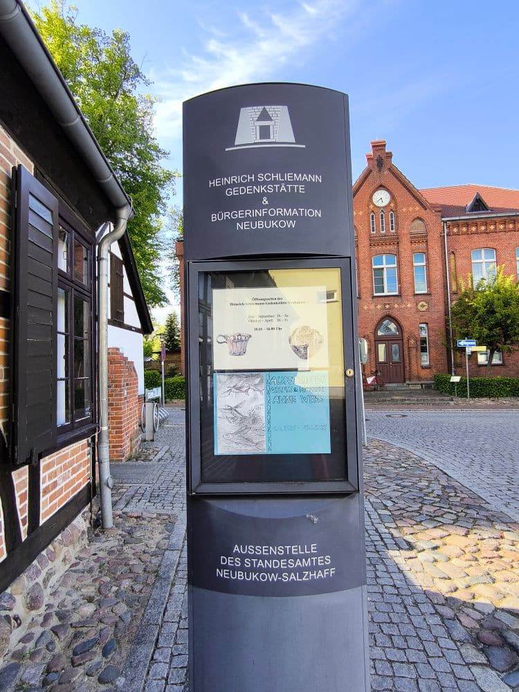 An einer Kopfsteinpflasterstraße in Neubukow steht ein hoher grauer Informationskiosk vor historischen Backsteinbauten. Er zeigt Aushänge und trägt die Aufschrift Heinrich Schliemann Gedenkstätte & Bürgerinformation Neubukow.