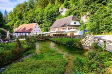 Der Blick auf einen klaren Bach, der neben zwei charmanten, rustikalen Häusern mit roten Ziegeldächern in Hayingen fließt, umgeben von üppig grünen Bäumen und einer Holzbrücke - perfekt für eine hochgehschätzt Wanderung Glastal an einem sonnigen Tag.