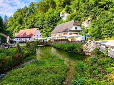 Der Blick auf einen klaren Bach, der neben zwei charmanten, rustikalen Häusern mit roten Ziegeldächern in Hayingen fließt, umgeben von üppig grünen Bäumen und einer Holzbrücke - perfekt für eine hochgehschätzt Wanderung Glastal an einem sonnigen Tag.