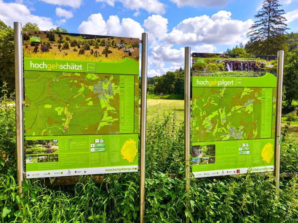 In der Nähe von Hayingen im Glastal stehen unter freiem Himmel im Grünen zwei grüne Wanderweg-Informationstafeln mit Karten und Texten für Ihre nächste Wanderung. Unter einem teilweise bewölkten blauen Himmel sind die Titel hochgehschätzt und hochgehpilgert zu lesen.