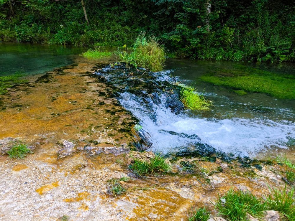 Ein kleiner Wasserfall fließt über mit grünem Moos und Gras bewachsene Felsen, umgeben von üppigem Grün und Bäumen bei Hayingen. Das klare Wasser sammelt sich unten und schafft einen ruhigen Ort entlang der Hochgehschätzt Wanderung Glastal.