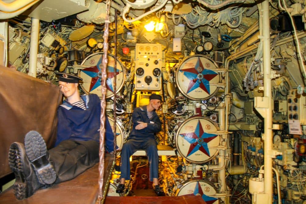 Zwei Matrosen in Uniform sitzen in einem überfüllten U-Boot-Kontrollraum, der an die Peenemünder Technik erinnert und mit Rohren, Drähten, Messgeräten, Maschinen und großen runden Luken gefüllt ist. Der überfüllte Raum ist hell erleuchtet.