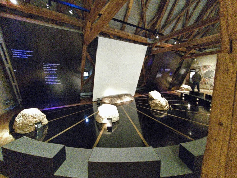Eine Ausstellung im Rieskratermuseum zeigt große Felsen in einem kreisförmigen Muster auf einem glänzend schwarzen Boden, an dessen Rand Bänke stehen. Holzbalken durchziehen die Decke, und Schautafeln beleuchten die Schwäbische Alb und die Geschichte Nördlingens.