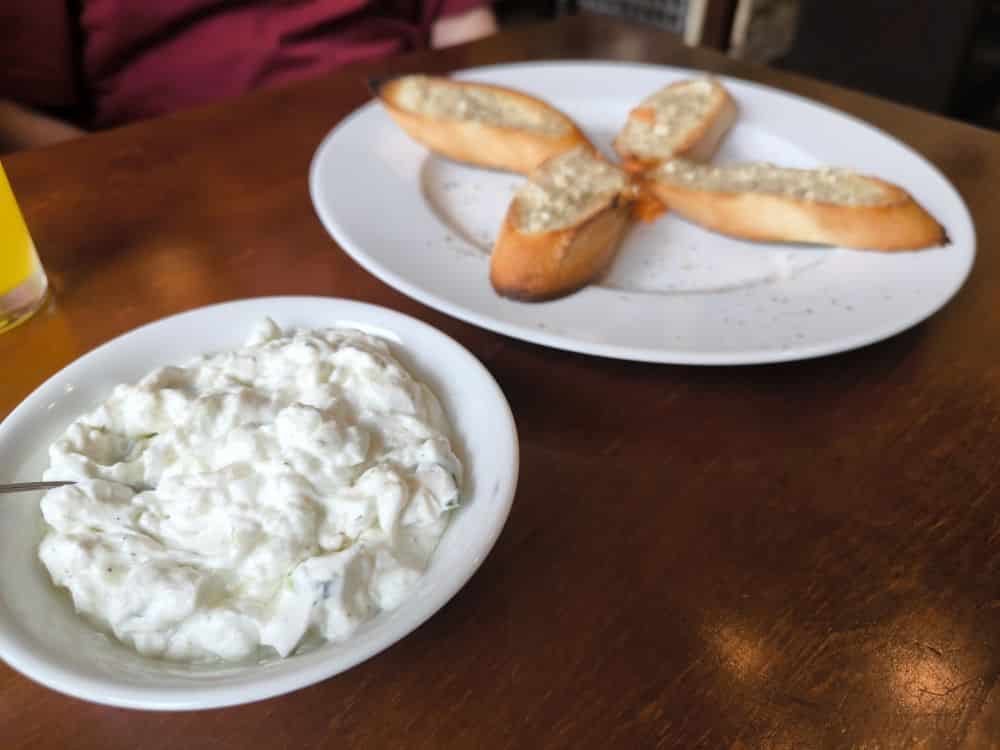 Eine Schale mit Tzatziki-Dip und ein Löffel stehen auf einem Holztisch neben einem Teller mit vier kreuzförmig angeordneten Stücken Knoblauchbrot - perfekt, um die vom Lieblingsrestaurant Rügen auf der schönen Insel Rügen inspirierten Aromen zu genießen.