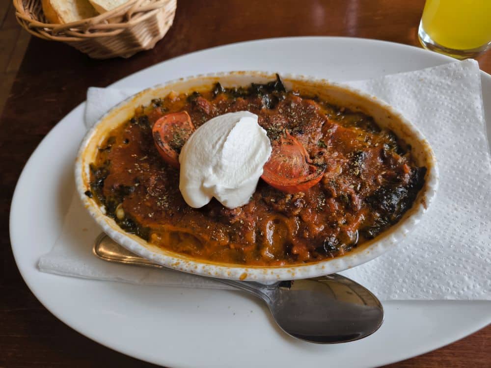 Ein Auflauf mit Tomatensoße, Hackfleisch, Spinat und Kräutern, übergossen mit Sahne und Tomatenscheiben - perfekt für den Genuss in einem der Lieblingsrestaurants auf Rügen, serviert auf einem weißen Teller mit einem Löffel und einem Brotkorb daneben.