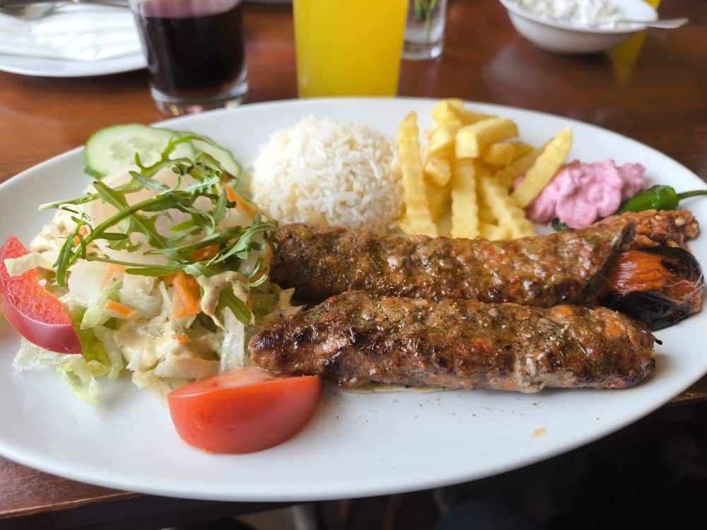 Ein Teller mit zwei gegrillten Spießen, Reis, Pommes frites, Salat mit Salat und Tomate, rosa Soße und Gurkenscheiben - perfekt zum Genießen in einem der Top-Lieblingsrestaurants Rügens. Rote und gelbe Getränke runden das Insel Rügen Menü ab.