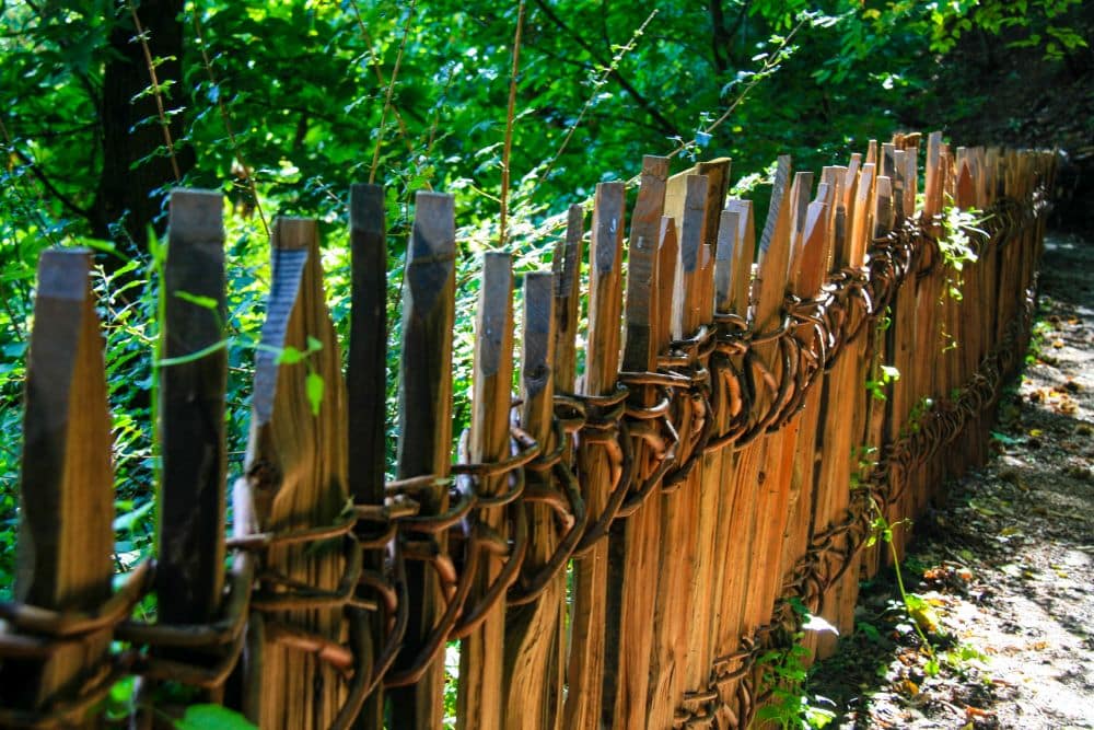 Ein rustikaler Holzzaun, der so genannte Brandiswaalweg, aus rauen, unebenen Pfählen, die mit Ästen verflochten sind, verläuft entlang eines schattigen Waldweges, umgeben von üppigem Grün und gedämpftem Sonnenlicht.