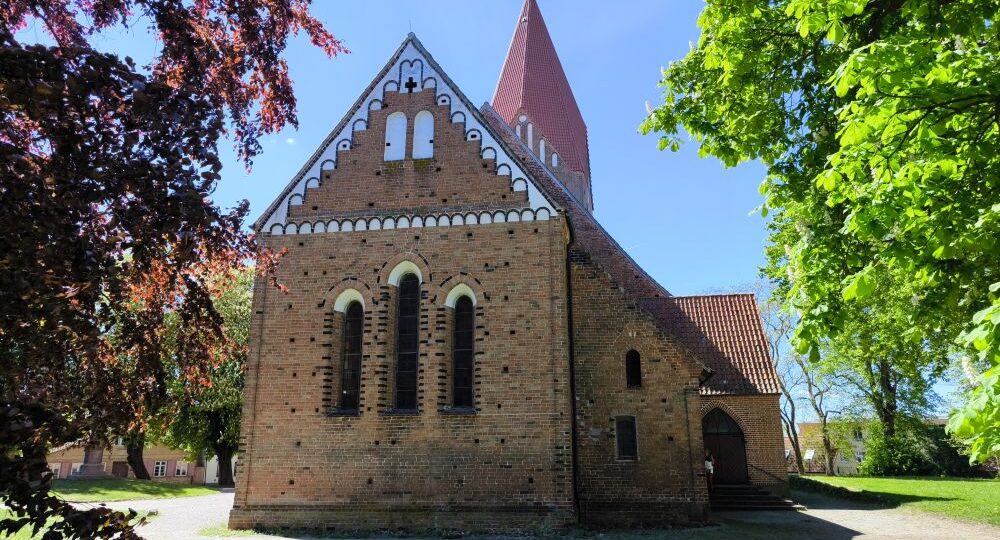 Eine Backsteinkirche mit steilem rotem Dach und Rundbogenfenstern steht an einem sonnigen Tag in Klütz an der Ostseeküste, umgeben von Bäumen und grünem Gras unter einem klaren blauen Himmel.
