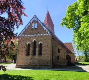 Eine Backsteinkirche mit steilem rotem Dach und Rundbogenfenstern steht an einem sonnigen Tag in Klütz an der Ostseeküste, umgeben von Bäumen und grünem Gras unter einem klaren blauen Himmel.