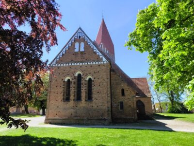 Eine Backsteinkirche mit steilem rotem Dach und Rundbogenfenstern steht an einem sonnigen Tag in Klütz an der Ostseeküste, umgeben von Bäumen und grünem Gras unter einem klaren blauen Himmel.