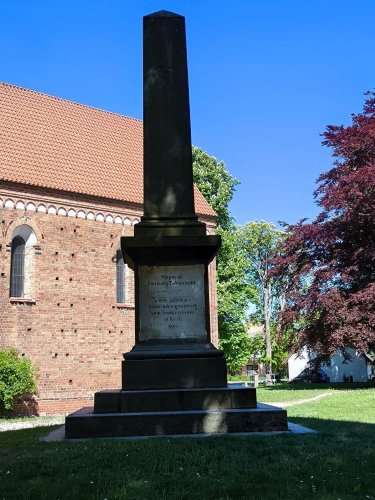 Ein hohes steinernes Obeliskdenkmal steht auf einem gestuften Sockel vor einem Backsteingebäude mit Rundbogenfenstern, umgeben von grünem Gras und Bäumen - dieses versteckte Juwel ziert die charmante Stadt Klütz an der Ostseeküste.