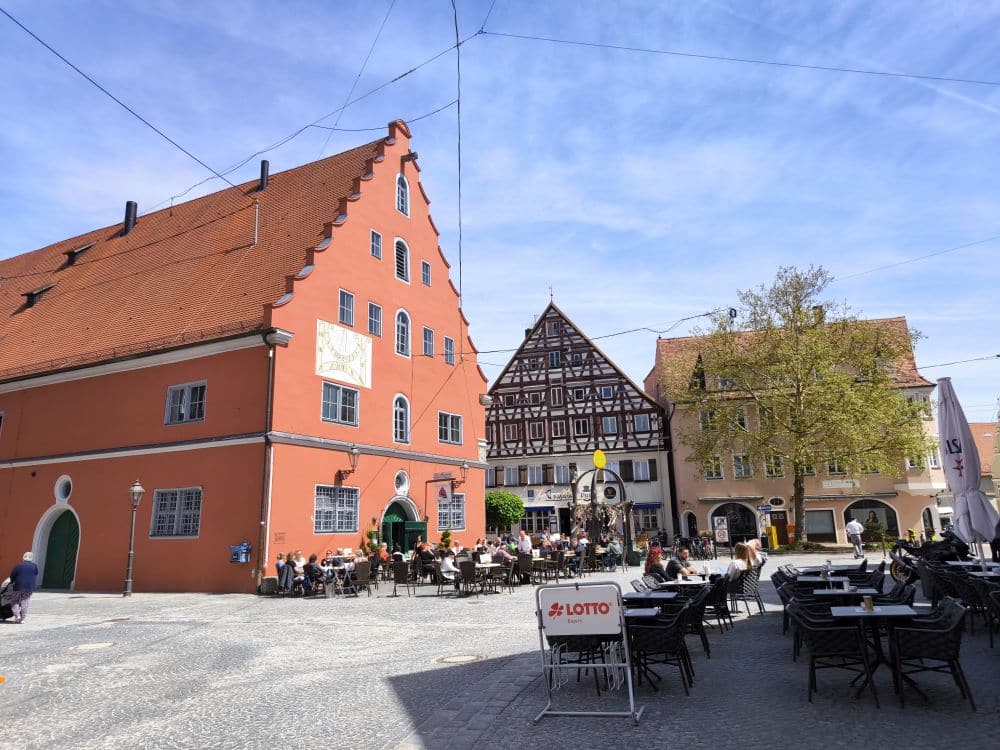 Ein sonniger Stadtplatz in Nördlingen mit Cafés, die im Freien sitzen, Menschen, die essen, und historischen Gebäuden mit roten, weißen und Fachwerkfassaden. Im Vordergrund ist ein Schild mit der Aufschrift LOTTO zu sehen.