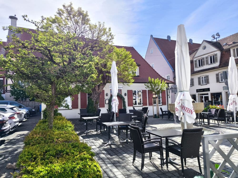 Außenterrasse eines europäischen Cafés in Nördlingen mit leeren Tischen und Stühlen, großen Sonnenschirmen, einem schattigen Baum und traditionellen Gebäuden mit roten Ziegeldächern im Hintergrund an einem sonnigen Tag.