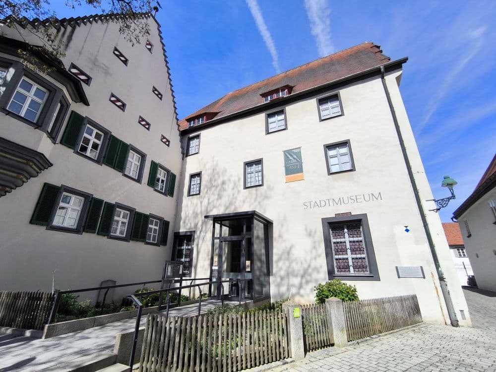 Ein weißes Gebäude mit rotem Ziegeldach und dem Schriftzug Stadtmuseum an der Wand steht unter einem blauen Wolkenhimmel in Nördlingen. Es hat mehrere Fenster, einen gläsernen Eingang und einen kleinen eingezäunten Gartenbereich davor.