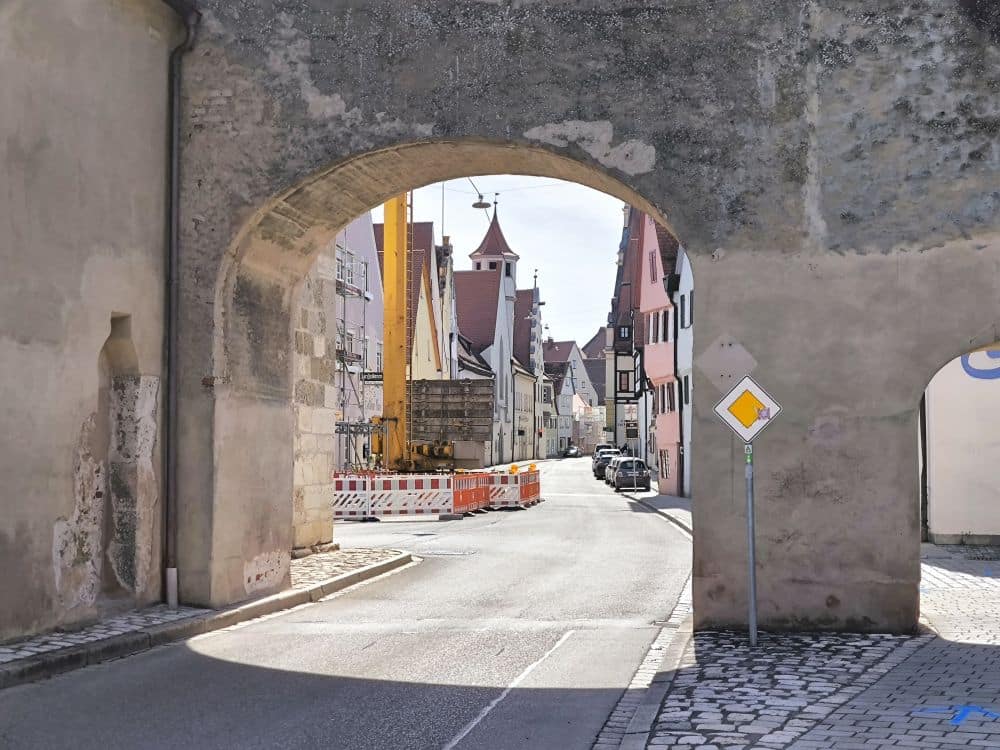 Ein Blick durch einen steinernen Torbogen auf eine schmale Straße in Nördlingen, in der Bauabsperrungen und Baumaschinen den Weg säumen; farbenfrohe historische Gebäude erhellen die Szene unter einem lebhaften Himmel.
