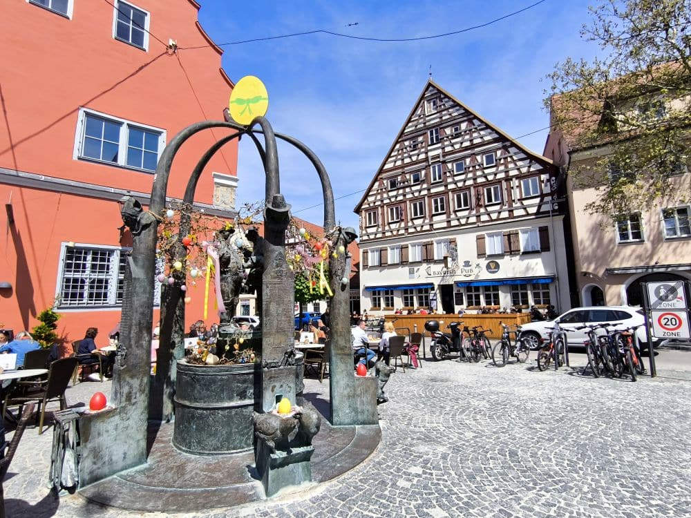 Ein festlicher, mit bunten Bändern und Eiern geschmückter Brunnen steht auf einem sonnigen Kopfsteinpflasterplatz in Nördlingen, umgeben von Sitzgelegenheiten im Freien, Fahrrädern und Fachwerkhäusern in dieser malerischen europäischen Stadt.