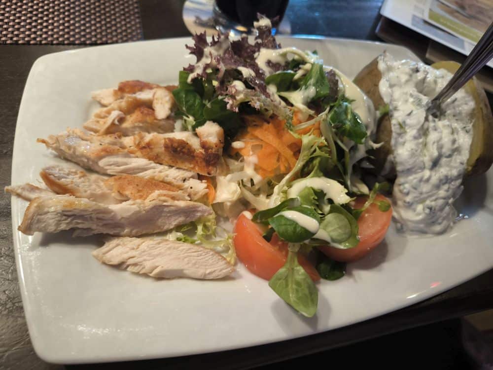 Ein Teller mit gegrilltem Hähnchenfleisch in Scheiben, ein gemischter Salat mit Salat, Tomate und Möhrenraspeln mit Dressing und eine Ofenkartoffel mit Sahnesoße und Kräutern - genau wie im Restaurant Rügen auf der schönen Insel Rügen.