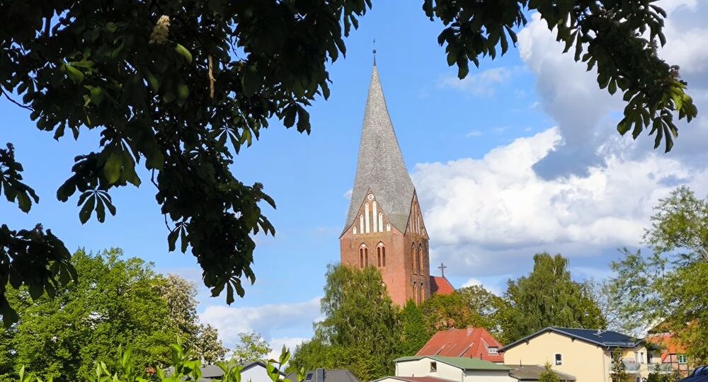 Ein hoher, spitzer Kirchturm in Neubukow erhebt sich über Bäumen und Häusern unter einem teilweise bewölkten blauen Himmel, eingerahmt von dunklen, belaubten Ästen im Vordergrund.