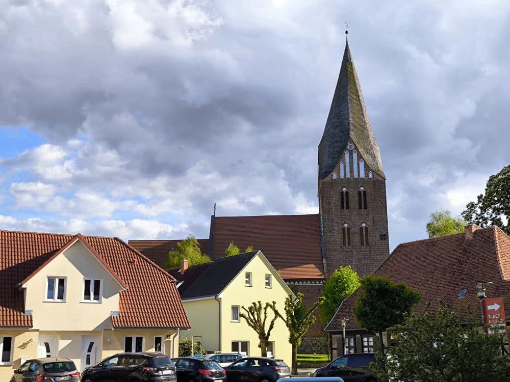 Eine Dorfszene in Neubukow mit mehreren Häusern im Vordergrund, geparkten Autos und einer hohen Kirche mit spitzem Kirchturm und Uhrenturm im Hintergrund, alles unter einem teilweise bewölkten Himmel.