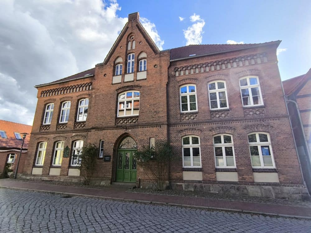 Ein großes, historisches Backsteingebäude in Neubukow mit Rundbogenfenstern, einer grünen Doppeltür und einem steilen Giebeldach steht an einer Kopfsteinpflasterstraße unter einem teilweise bewölkten Himmel.