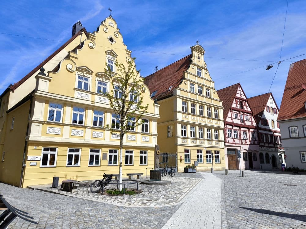 Ein sonniger Stadtplatz in Nördlingen mit Kopfsteinpflaster, verzierten gelben und roten Fachwerkhäusern, einem schattigen Baum, Bänken und abgestellten Fahrrädern unter strahlend blauem Himmel.
