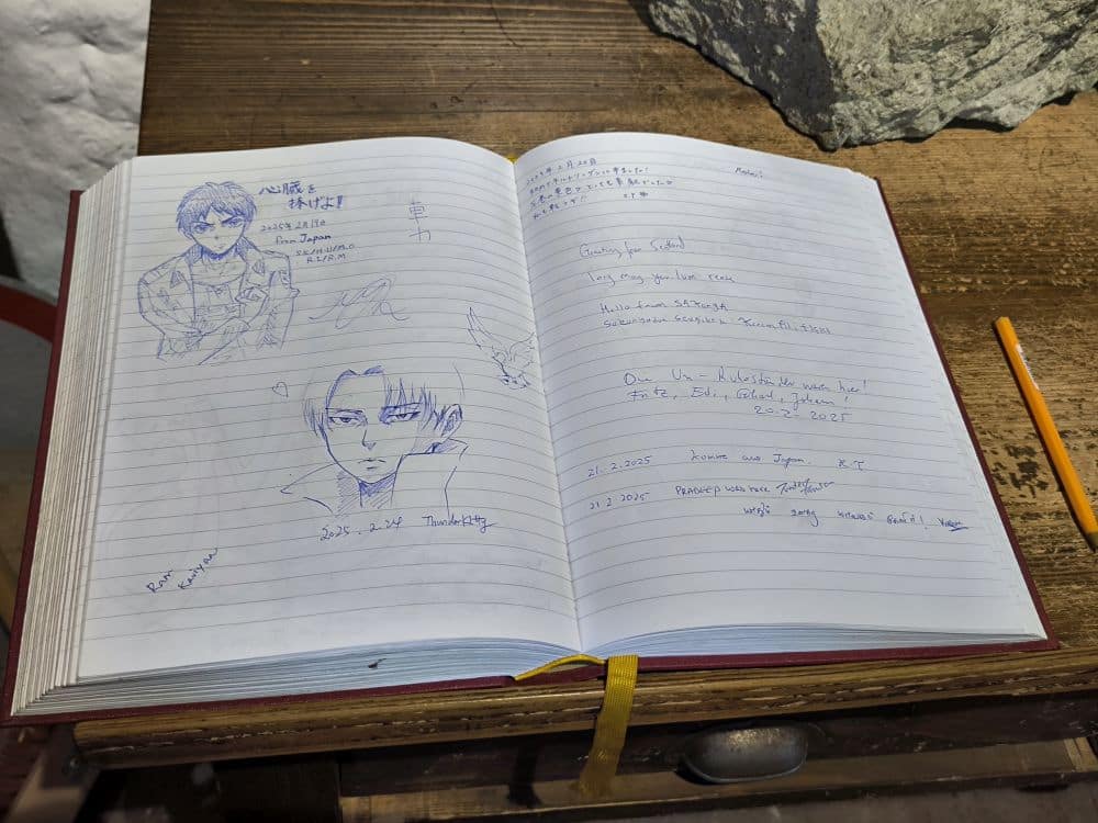 Ein aufgeschlagenes Gästebuch auf einem Holztisch in Nördlingen zeigt handschriftliche Notizen in Englisch und einer anderen Sprache, Bleistiftskizzen von zwei männlichen Gesichtern im Anime-Stil und einen gelben Bleistift, der neben dem Buch liegt.