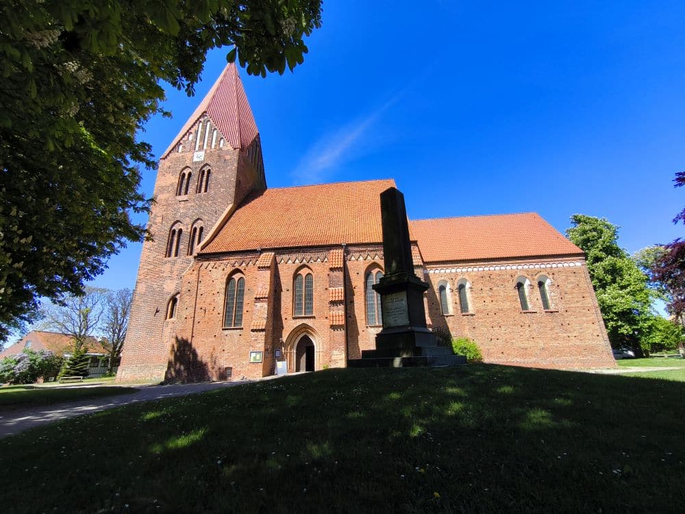 In Klütz, einem versteckten Juwel an der Ostseeküste, steht eine Backsteinkirche mit hohem Kirchturm und rotem Ziegeldach auf einer Wiese unter strahlend blauem Himmel; im Vordergrund steht ein steinernes Denkmal, das teilweise von Bäumen beschattet wird.
