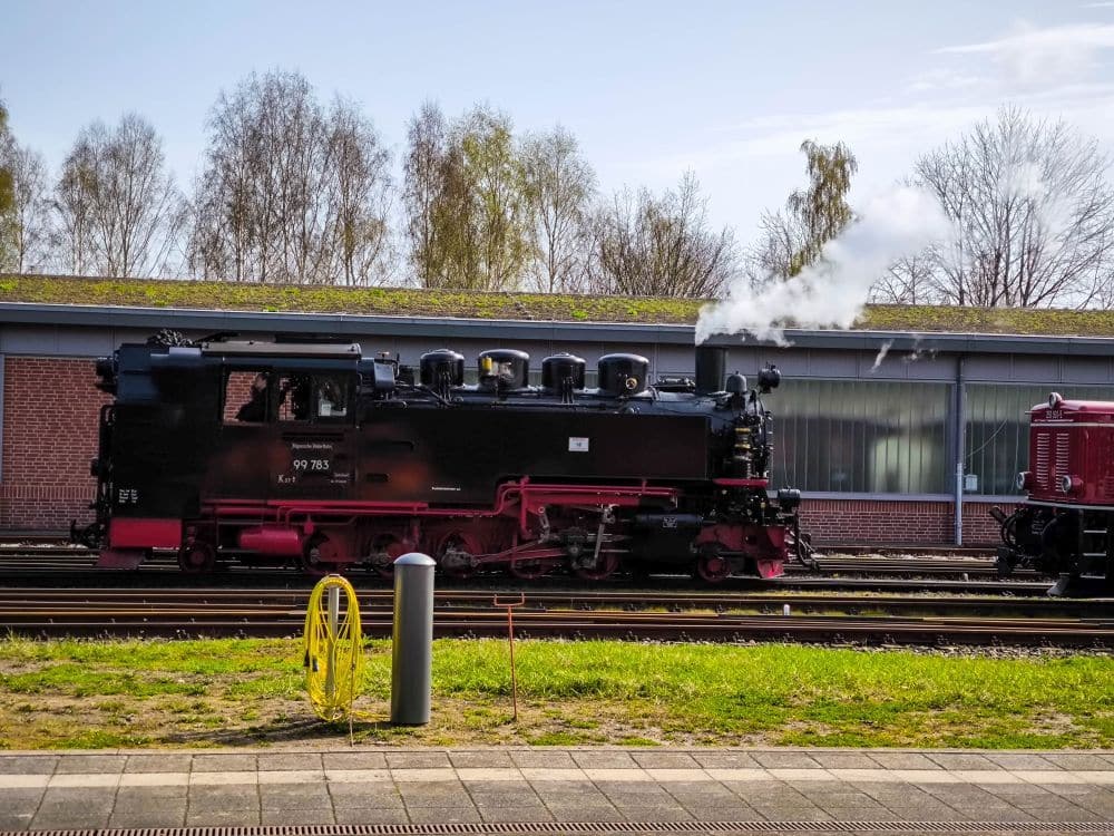 Eine schwarz-rote Dampflokomotive des Rasenden Rolands stößt weißen Dampf auf den Gleisen neben einem Backsteingebäude aus, mit Bäumen und blauem Himmel im Hintergrund. Im Vordergrund sind eine gelbe Bahnschranke und grünes Gras auf Rügen zu sehen.