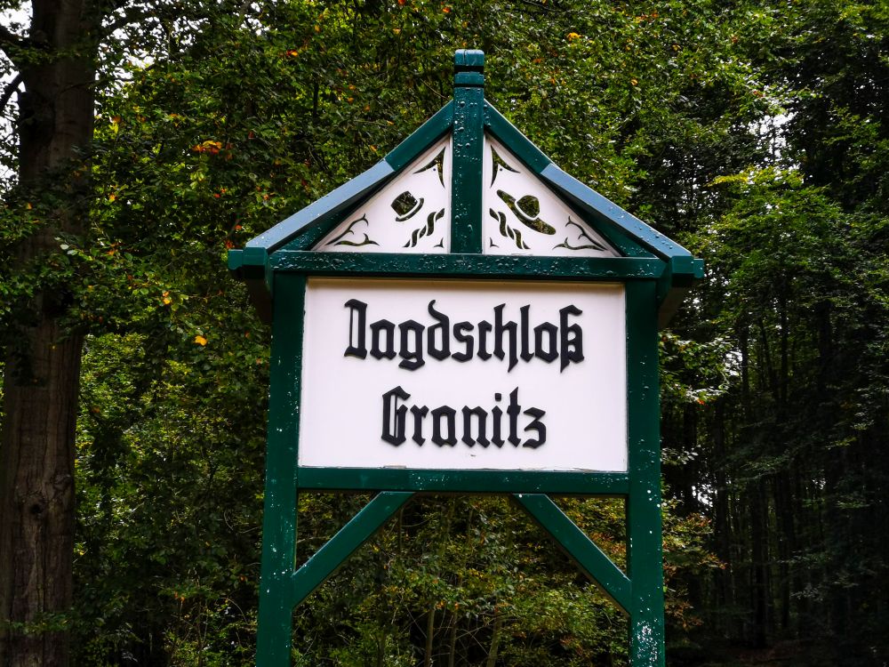 Auf einem grün umrahmten Schild mit dekorativen Elementen steht in schwarzer gotischer Schrift Jagdschloss Granitz vor dem Hintergrund eines dichten, belaubten Rügener Waldes, an dem der historische Rasende Roland oft vorbeidampft.