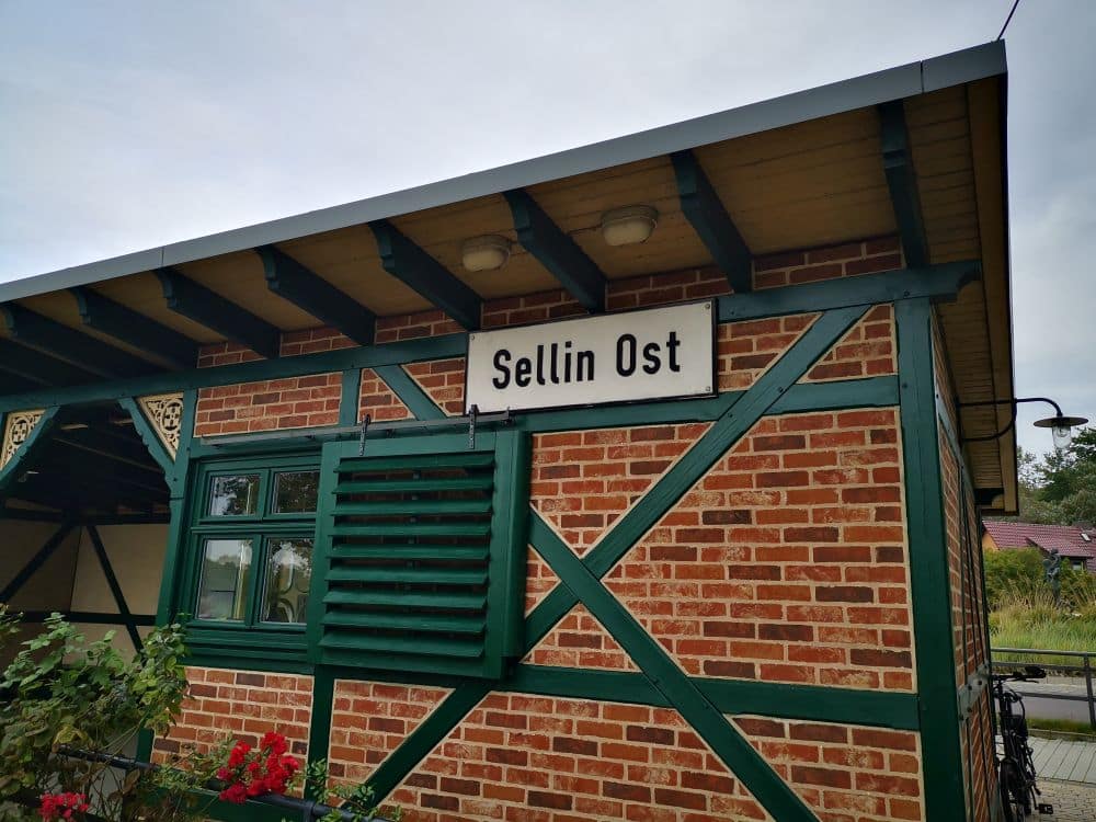 Ein Backsteingebäude mit grünen Holzbalken trägt unter seinem Dach ein Schild mit der Aufschrift Sellin Ost - eine charmante Haltestelle des berühmten Rasenden Rolands auf Rügen. Vor dem Gebäude gibt es ein Fenster mit grünen Fensterläden und einige Pflanzen.