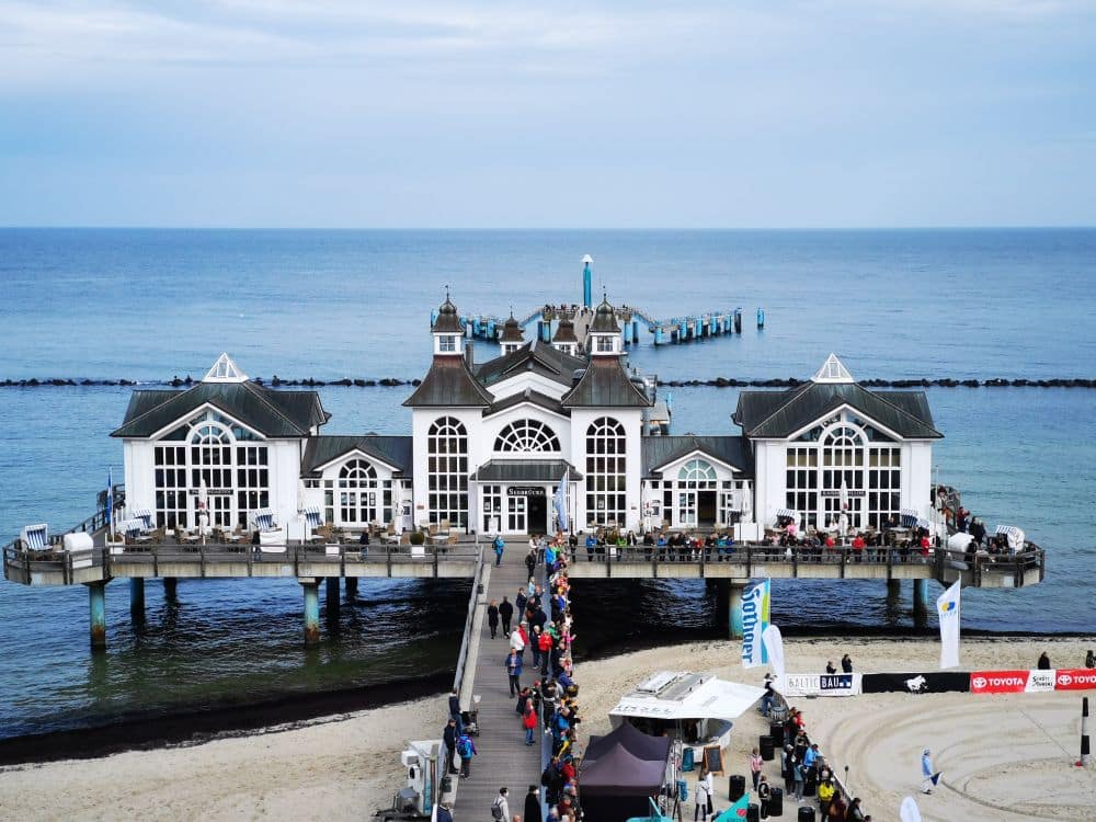 Eine malerische Seebrücke mit einem weißen, verzierten Gebäude erstreckt sich über das blaue Meer auf Rügen, flankiert von Sandstränden. Bei teilweise bewölktem Himmel spazieren die Menschen an der Seebrücke entlang und versammeln sich am Ufer.