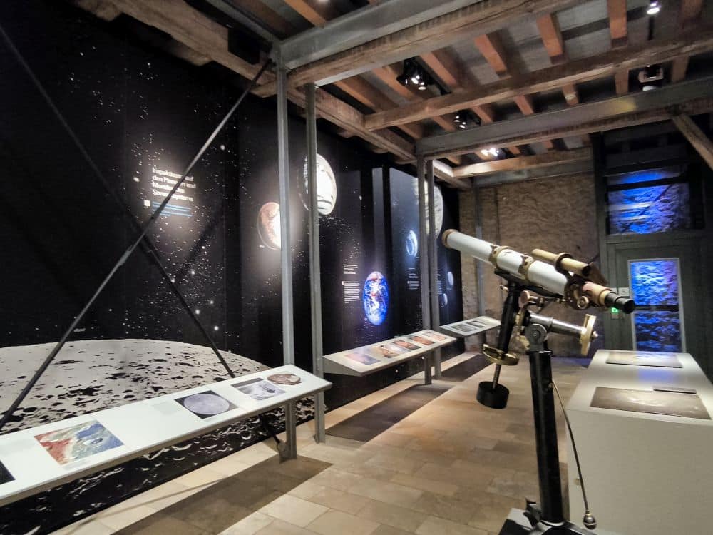 Eine Museumsausstellung im Rieskratermuseum in Nördlingen zeigt Planetenbilder an einer schwarzen Wand, Informationstafeln auf einer Theke und ein Messingfernrohr unter einer Holzdecke. Blaues Licht beleuchtet die Steinwand der Schwäbischen Alb dahinter.