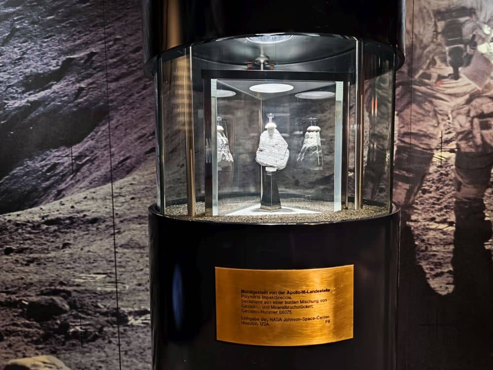 Eine Mondgesteinsprobe von der Apollo 16-Mission wird im Rieskratermuseum in Nördlingen in einer Glasvitrine ausgestellt. Darüber befinden sich Scheinwerfer, darunter eine goldene Plakette und im Hintergrund ein Bild der Mondoberfläche mit Astronautenfiguren.
