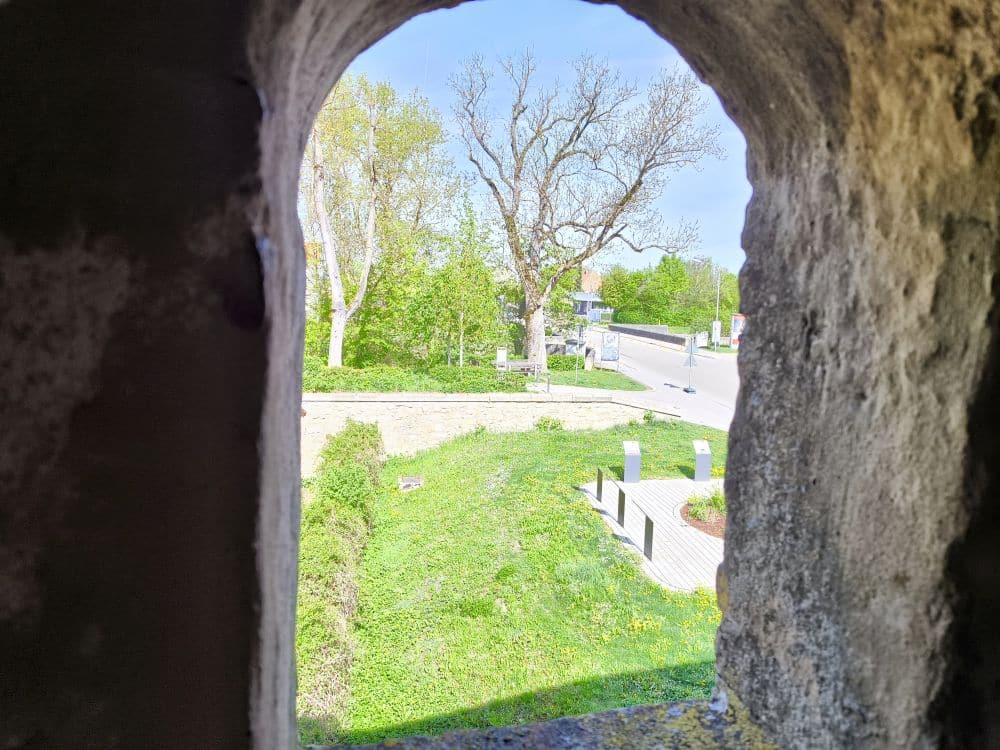 Der Blick durch ein steinernes Fenster in Nördlingen zeigt eine Wiese mit Bäumen, eine Steinmauer und eine nahe gelegene Straße mit Bürgersteigen. Es ist ein heller, sonniger Tag mit strahlend blauem Himmel und üppigem Grün.