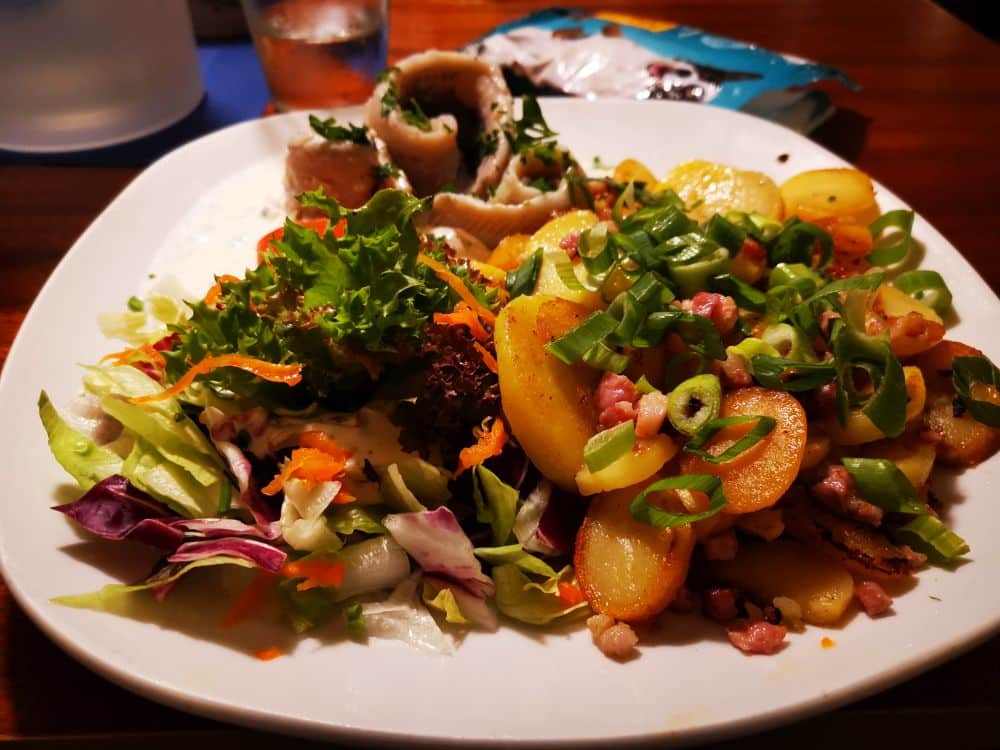 Ein Teller mit frischem grünen Salat, Bratkartoffeln mit Frühlingszwiebeln und Speck und gerollten Fischscheiben, garniert mit Kräutern - so wie in einem der Lieblingsrestaurants Rügen auf der Insel Rügen.