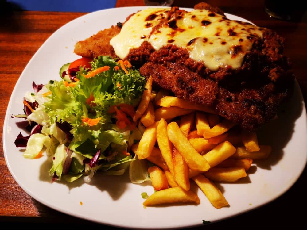 Ein Teller mit paniertem Schnitzel, überbacken mit geschmolzenem Käse und Fleischsauce, serviert mit goldgelben Pommes frites und einem frischen gemischten Salat - perfekt zum Genießen in einem der Lieblingsrestaurants auf der schönen Insel Rügen.
