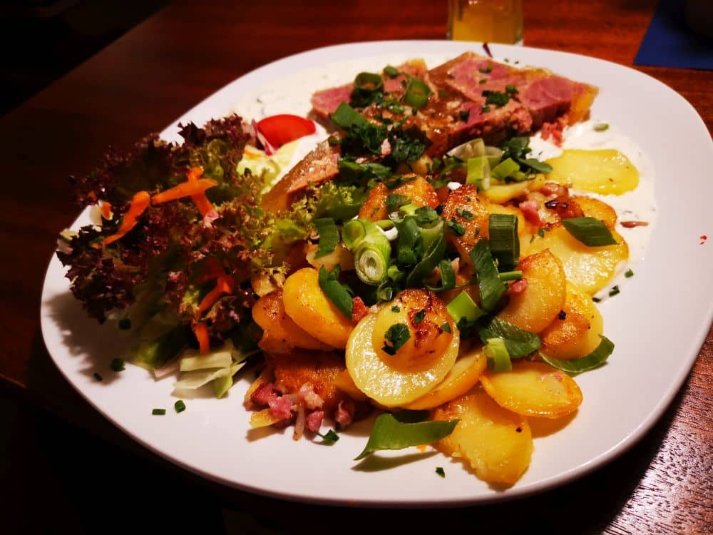 Ein weißer Teller mit in Scheiben geschnittenen Kartoffeln, garniert mit grünen Zwiebeln, ein frischer Blattsalat mit geraspelten Möhren und saftige Fleischscheiben, alles serviert auf einem Holztisch - ein Vorgeschmack auf das Restaurant Rügen auf der schönen Insel Rügen.